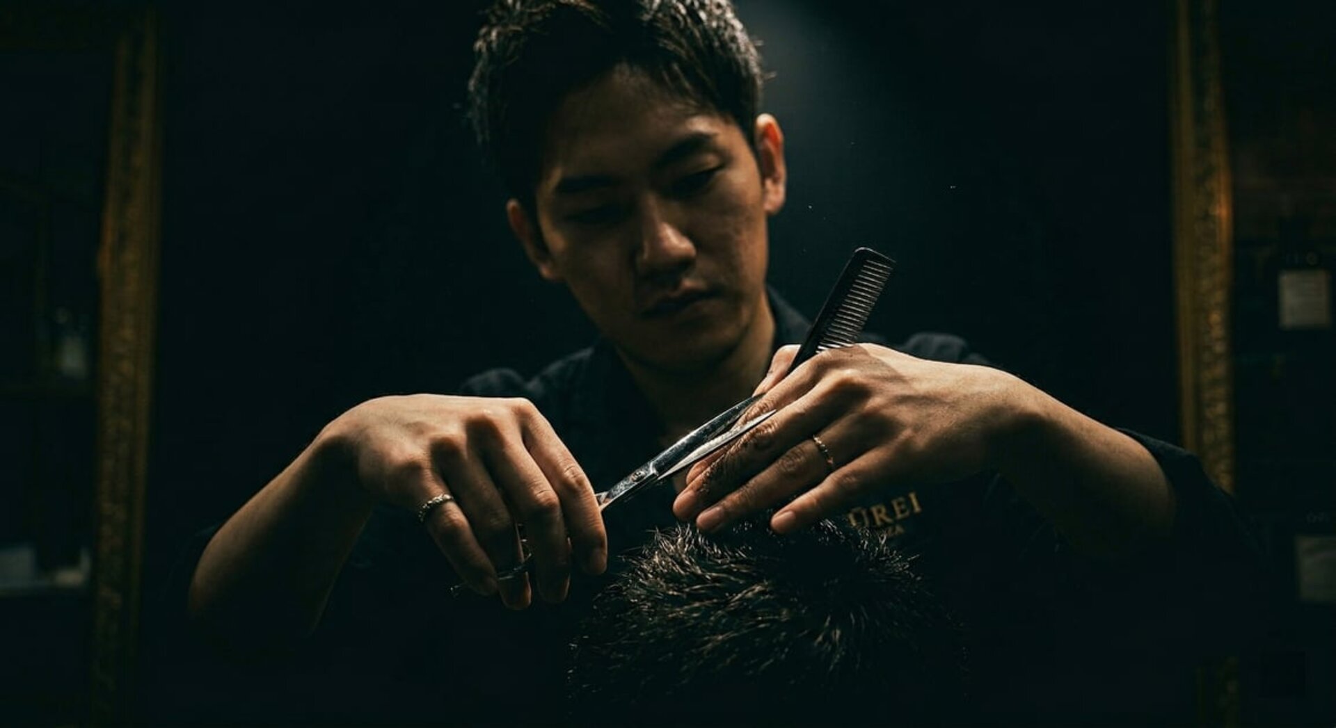 Professional barber hands with scissors performing precision cutting technique in dark moody lighting