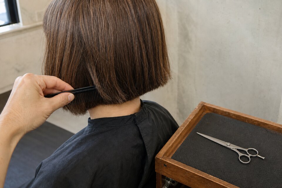 A stylist uses a matte comb to visually check the fall of a completed blunt bob while a fully closed generic shear rests separately on a trolley.