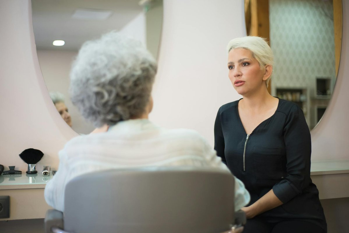 En stylist med kort platinablondt hår sitter foran et rundt salongspeil overfor en eldre klient med kort grått hår.