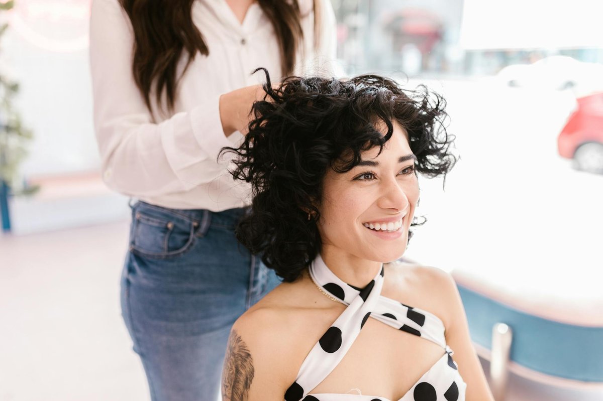En person med skulderlangt krøllet hår sidder i en lys salon, mens en stylist arbejder i nærheden.