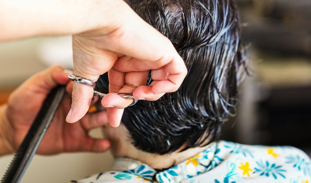 A stylist cuts the nape of short, visibly wet dark hair with a silver shear while holding a black comb.