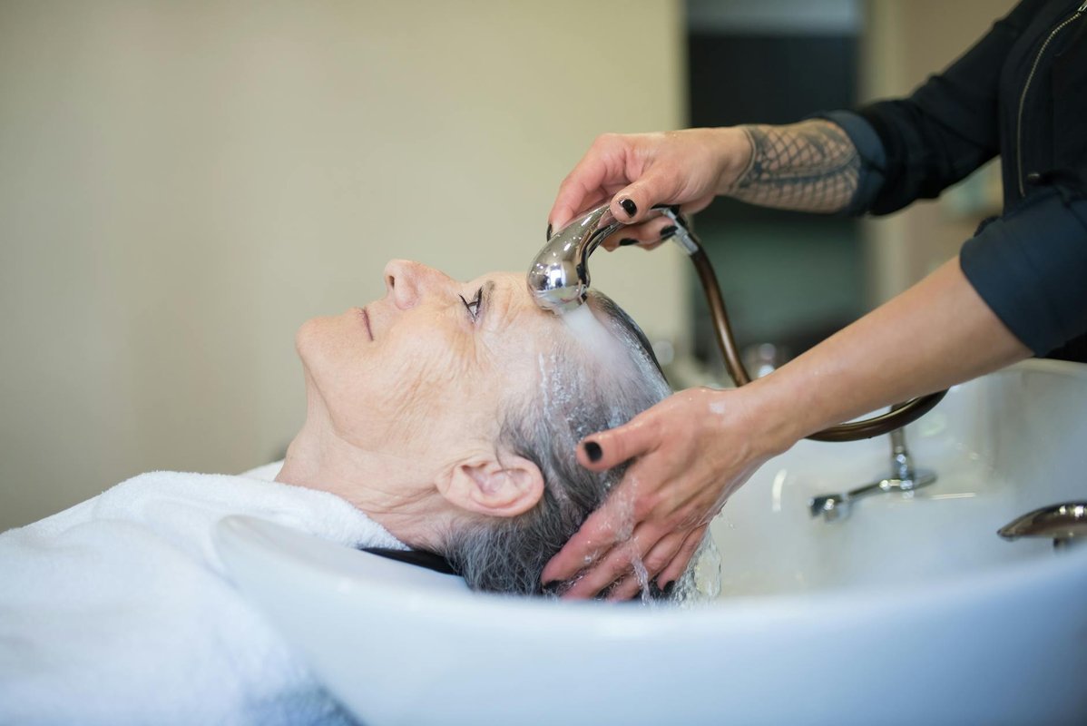 An older woman reclines at a basin while a salon worker washes her hair.