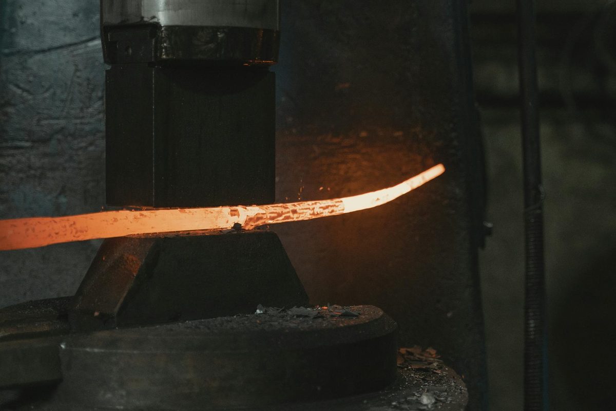 A glowing metal bar is shaped under an industrial forming machine; the photograph is general hot-metal context, not a documented shear factory.