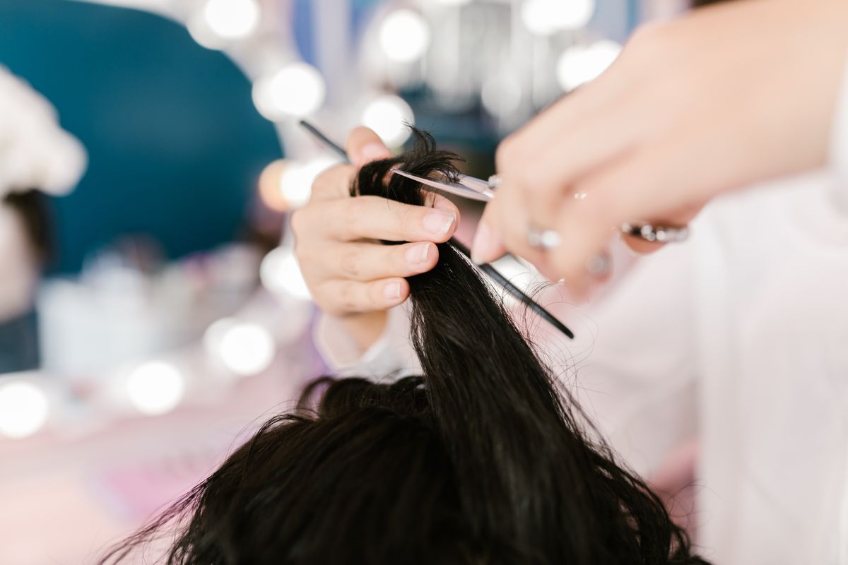 A stylist holds a cutting shear and comb while working on a dark hair section.