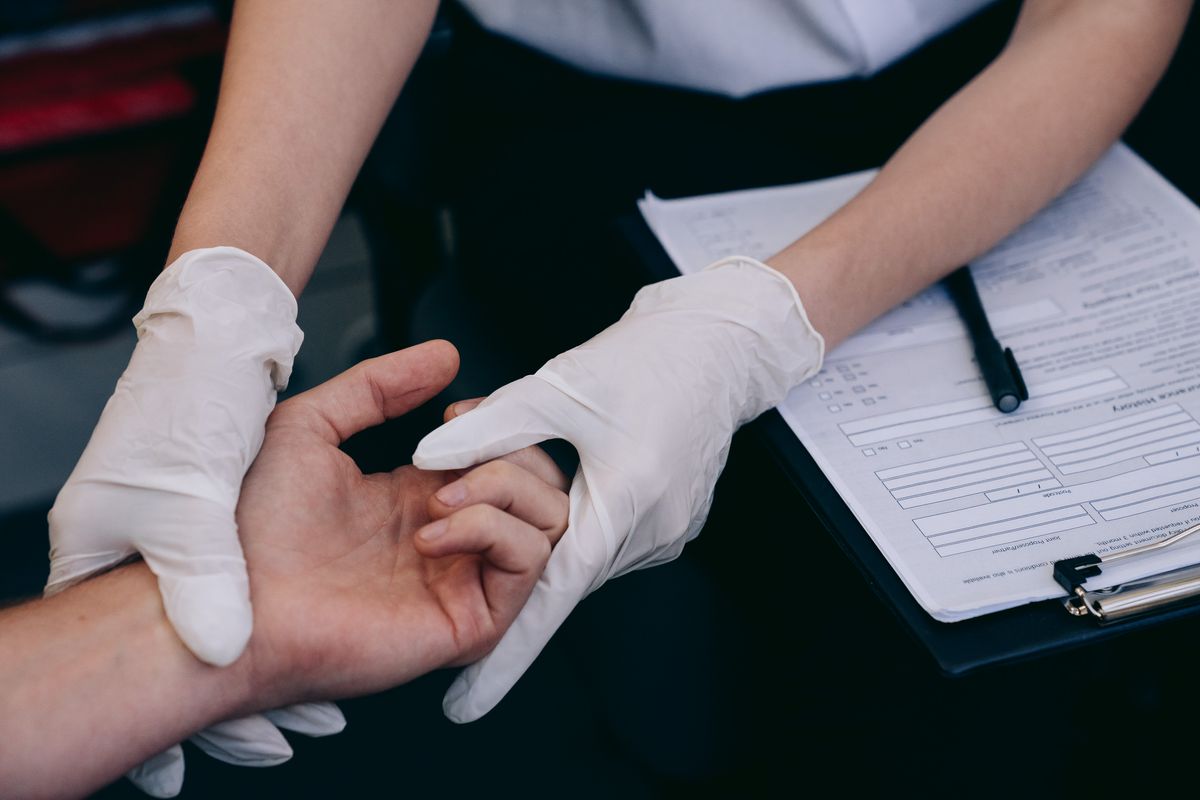 A gloved healthcare worker examines a person's hand beside a medical clipboard.
