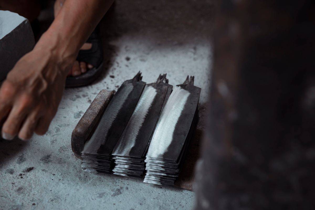 Rows of unfinished metal blade blanks lie on a workshop floor beside a worker.