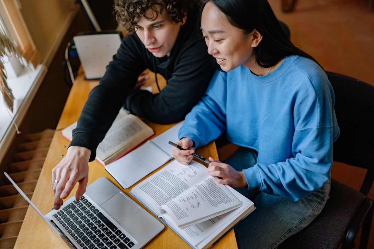 Two students work together with a laptop, notebook, and open reference books.