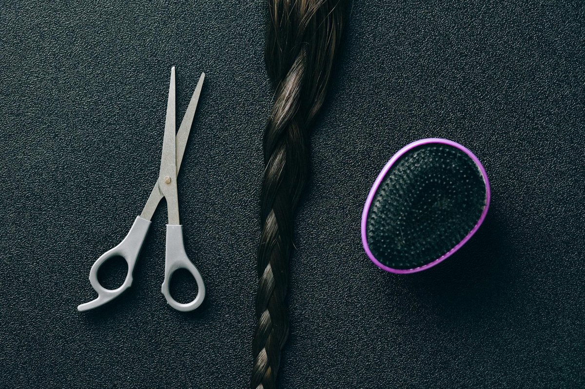 A cutting shear, braided hair sample, and oval brush on a dark surface.