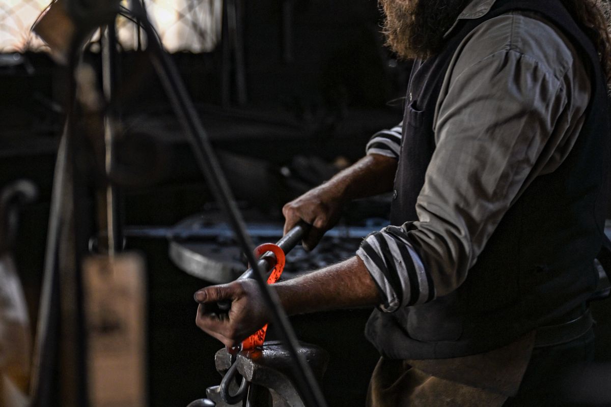 A blacksmith handles glowing metal with tongs in a dark traditional workshop.