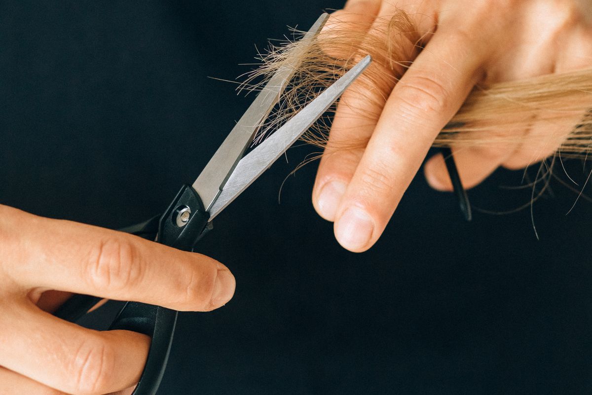 A close view shows a stylist trimming blonde hair with a metal cutting shear.