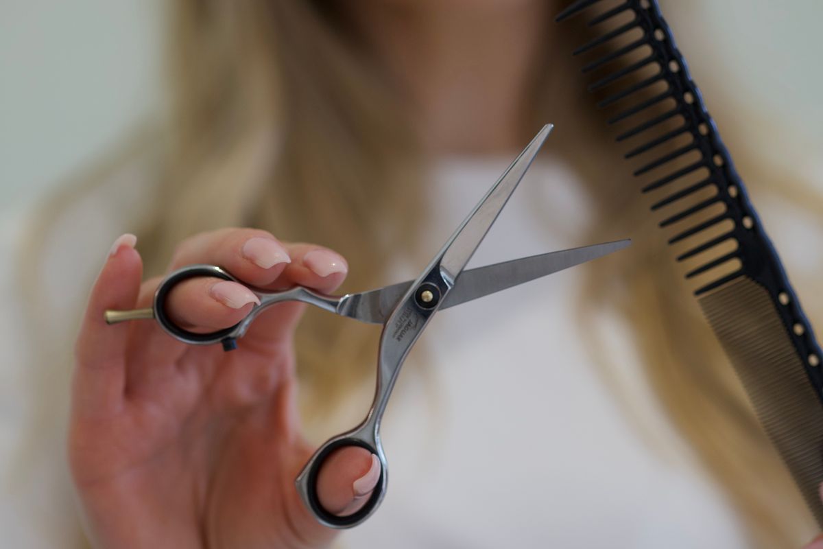 A stylist holds an open silver cutting shear and a black comb in a salon.