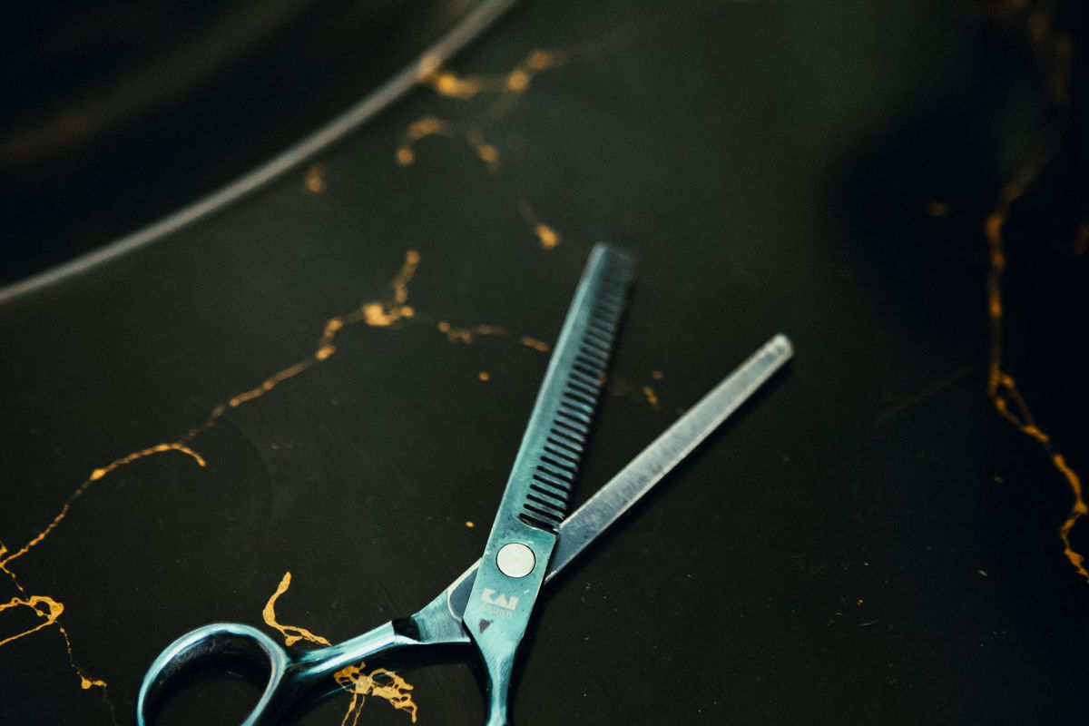 A professional thinning shear with one toothed blade lies beside a slim comb on a black surface.